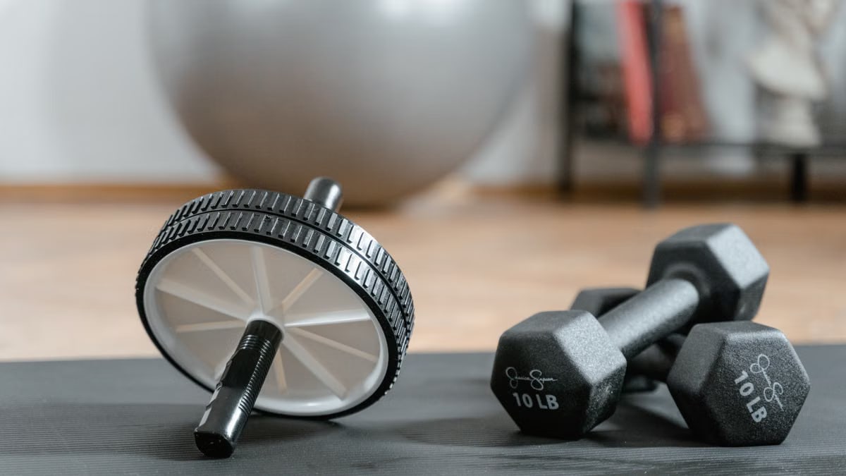 Close-up of ab wheel and dumbbells on a mat in a home gym setting.